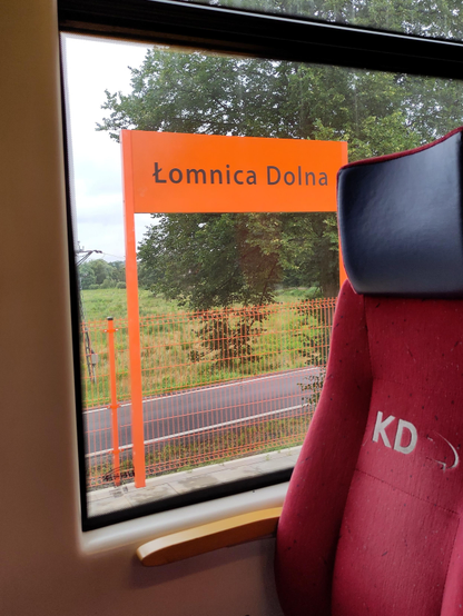 Widok przez okno pociągu Kolei Dolnośląskich, co można poznać na napisie na fotelu. Za oknem tabliczka z nazwą stacji Łomnica Dolna. Zarówno tablica jak i płot za nią są koloru jaskrawo pomarańczowego.