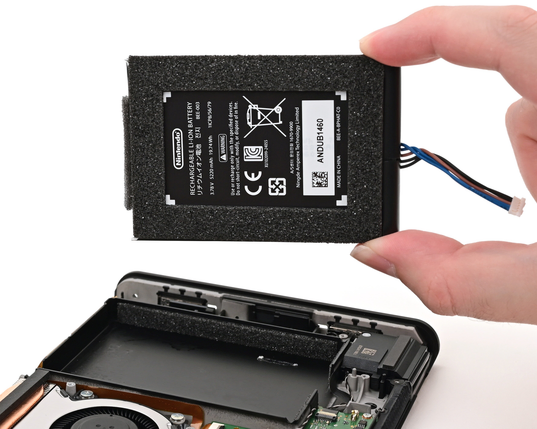 A hand holds a Nintendo Switch 2 lithium-ion battery above the open handheld console during a battery replacement. The console's internal components are visible below.