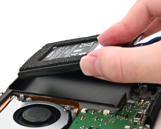 A hand lowers a Nintendo Switch 2 battery into the open console during reassembly. The battery is positioned above the battery compartment, with the console's motherboard visible below.